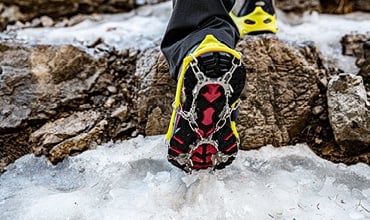 Nahaufnahme einer Person, die auf eisbedeckten Felsen wandert. Sie trägt leuchtend gelbe und schwarze Schuhe, die mit Metallsteigeisen für den Halt ausgestattet sind. Die Szene betont Winteraktivitäten im Freien.